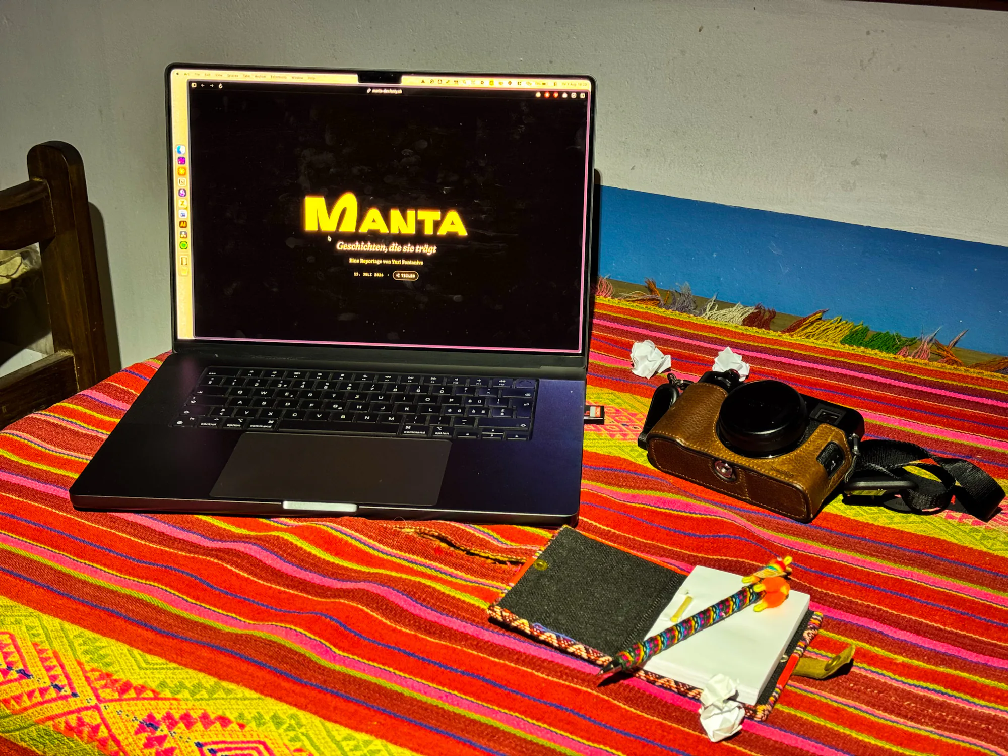 Eine Kamera, ein Notizbuch und ein Laptop liegen auf einer farbig gestreiften Manta.