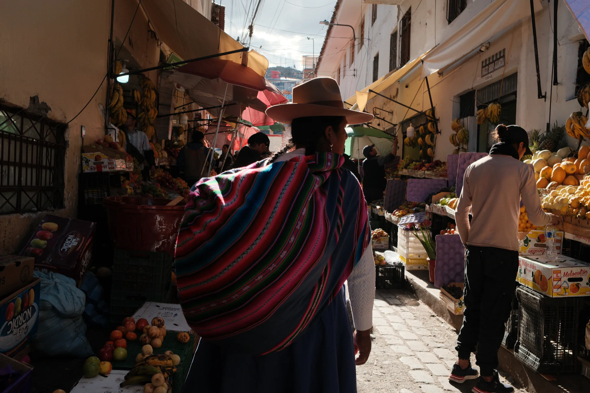 Eine Frau geht mit einer grossen gestreiften Manta auf dem Rücken durch einen Markt in Cusco.